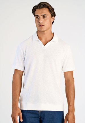 Jonge man die een witte polo met korte mouwen en een blauwe spijkerbroek draagt, staand tegen een effen lichte achtergrond.