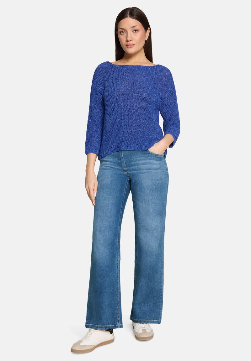 Femme portant un pull texturé bleu royal, un jean large bleu clair et des baskets blanches, debout avec une main dans la poche.