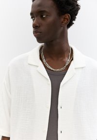 Camicia bianca a maniche corte con texture, con chiusura a bottoni e colletto, indossata sopra una canotta grigia a costine, completata da collane a catena sovrapposte.