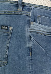 Blue denim jeans with yellow stitching, a label on the back pocket, and a small front pocket design, featuring a slightly faded texture.