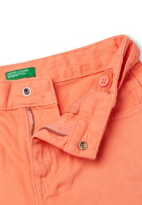 Korálové džínové nohavice z bavlny se zapínáním na zip a knoflík, s viditelnými švy a zelenou etiketou "United Colors of Benetton".