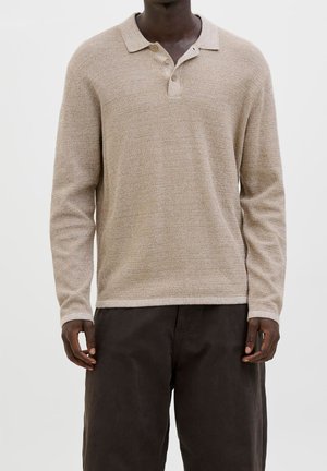 Homme portant un polo à manches longues beige texturé avec des boutons et un pantalon ample marron foncé sur un fond uni.