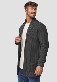 Cardigan grigio scuro con tessuto lavorato a maglia, apertura frontale, polsini a coste e una tasca frontale, indossato sopra una camicia chiara.