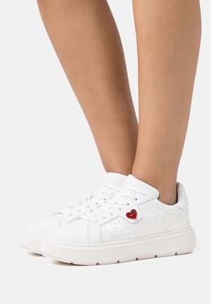 Zapatillas de cuero blanco con suela plana, que presentan un diseño de cordones, herrajes en forma de corazón y texto embossado en el lateral.