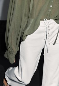 Blusa trasparente di colore verde oliva con maniche ampie abbinata a pantaloni bianchi a vita alta con lacci frontali e texture sottile.
