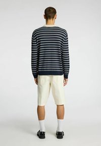 Maglione a maniche lunghe navy e bianche a righe con polsini e orlo a coste, abbinato a pantaloni corti beige chiaro, calze bianche e scarpe nere.