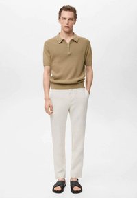 Beige poloshirt met korte mouwen en een ritssluiting bij de kraag, gecombineerd met lichtbeige broeken en zwarte sandalen. Gladde textuur overal.