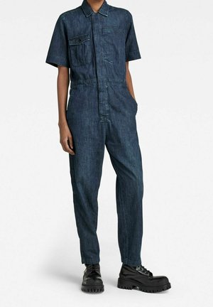 Granatowy kombinezon z denimu z krótkimi rękawami, kieszenią na piersi i zamkiem z przodu. Luźny krój z kieszeniami po bokach oraz solidne czarne buty.