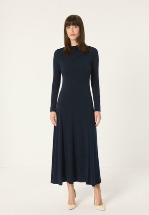 Žena stojaca v dlhých tmavomodrých šatách s dlhým rukávom po členky a béžových podpätkoch na jednofarebnom svetlom pozadí.