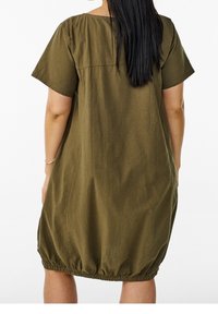 Mujer con cabello largo y negro que lleva un vestido suelto de color verde oliva con mangas cortas y dobladillo fruncido, de pie de espaldas al espectador.