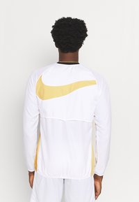 Mann von hinten in weißer Sportkleidung mit einem großen goldenen Swoosh-Logo auf dem Rücken und passenden Shorts vor einem einfarbigen Hintergrund.