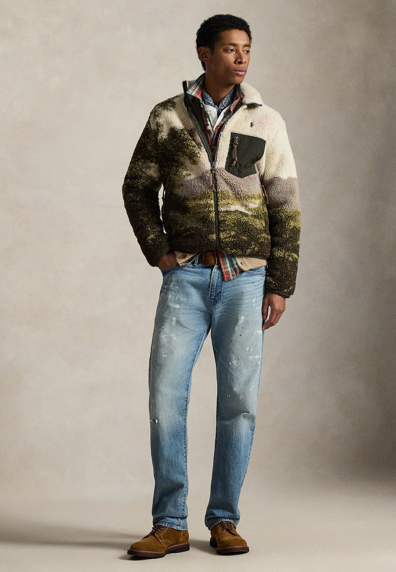 Giacca sfocata e multicolore con chiusura frontale a zip, dettaglio tasca verde, abbinata a jeans azzurri consumati e stivali marroni con lacci.