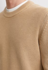 Beige Strickpullover mit gerippter Textur, Rundhalsausschnitt und subtil gleichmäßigen Mustern. Zeigt Nahaufnahmen der Stoffdetails und des Designs.