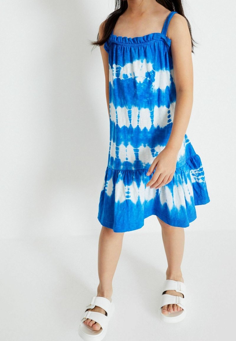 Enfant portant une robe sans manches tie-dye bleue et blanche et des sandales blanches, debout sur un fond blanc uni.