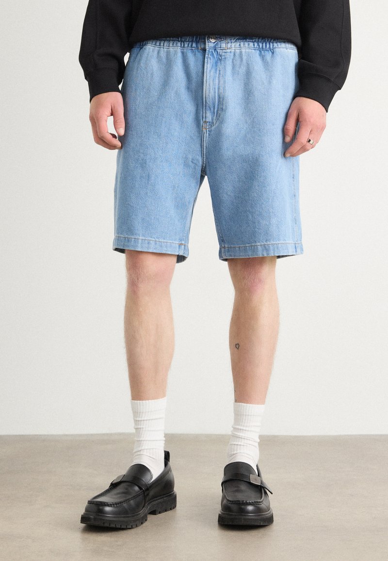 Shorts in denim azzurro chiaro con vestibilità rilassata, orli risvoltati e chiusura con bottone. Abbinati a mocassini neri e calze bianche a coste.