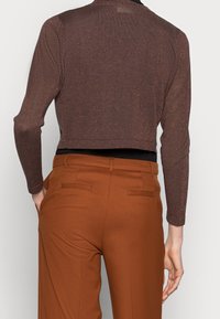 Osoba ubrana w błyszczący brązowy crop top z długim rękawem i wysokim stanem w kolorze rdzy, z rękami spoczywającymi blisko kieszeni, odwrócona plecami.