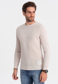 Pull en tricot beige clair avec des manches longues, des poignets et un ourlet côtelés, un motif texturé et une étiquette sur le côté gauche en bas. Associé avec un jean bleu.