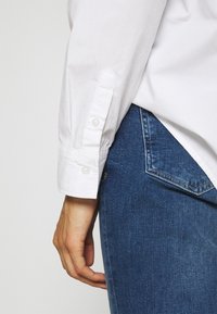 Weißes Langarmhemd mit Knopfmanschette, kombiniert mit dunkelblauen Jeans aus Denim, die eine dekorative Gesäßtasche und eine subtile Textur aufweisen.