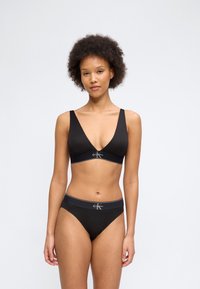 Γυναίκα με σγουρά μαλλιά που φοράει μαύρο bralette Calvin Klein και ταίριαστο εσώρουχο, στέκεται μπροστά σε ένα απλό ανοιχτόχρωμο φόντο.