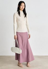 Haut crème à manches longues avec un col froncé, associé à une jupe maxi rayée rose et violette et des ballerines tissées beiges, tenant un sac blanc.