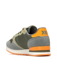 El zapato presenta una parte superior de ante y malla en tonos verde y gris, con acentos naranjas en el talón y el logo, y una suela blanca y naranja. Diseño redondeado.