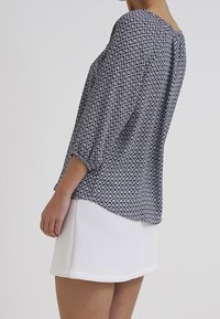Blouse à manches longues en motif géométrique bleu foncé et blanc, en tissu léger, coupe ample, légèrement drapée, associée à une mini-jupe blanche structurée.
