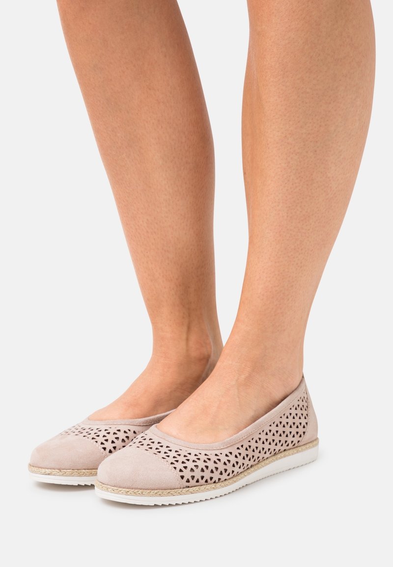 Jambes portant des chaussures plates rose clair avec des motifs triangulaires découpés et des semelles en caoutchouc blanches, debout sur un fond clair uni.