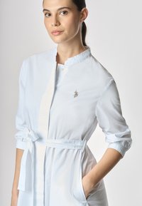 Abito camicia bianco in tessuto leggero con colletto, vita regolabile, maniche lunghe con polsini arricciati e un piccolo dettaglio ricamato.
