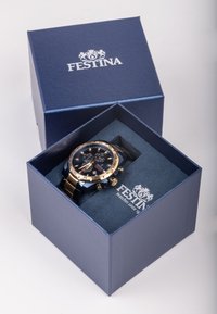 Festina laikrodis tamsiai mėlyname dėžutėje. Laikrodis turi tamsų ciferblatą, auksinius akcentus, chronografo funkcijas ir metalinę apyrankę. Dėžutė turi minkštą vidų.
