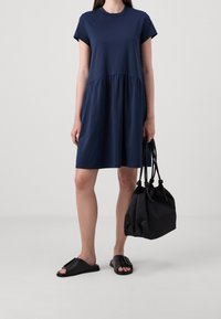 Vrouw in een marineblauwe jurk met korte mouwen en een geplooide taille, die een zwarte trekkoordtas vasthoudt en zwarte slipper-sandalen draagt.