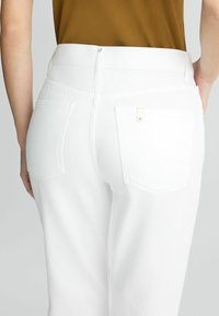 Pantalones de mezclilla blancos con una cintura alta, dos bolsillos traseros con herrajes dorados y una textura suave. No se observan embellishments adicionales.