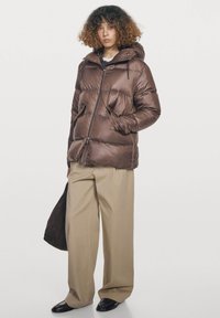 Person står iført en skinnende brun dunjakke med hætte, beige bukser med vide ben, sorte sko og holder en mørkebrun taske.