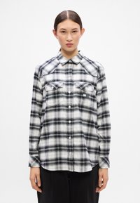 Camisa de franela de cuadros en blanco y negro con cuello abotonado, dos bolsillos en el pecho y mangas largas. Textura suave y diseño casual.