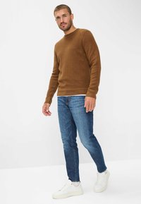 Bruine ribgebreide trui, blauwe jeans en witte sneakers. De trui heeft een ronde hals; de jeans hebben een slim fit met een standaard taillehoogte. Schoon, minimalistisch ontwerp.