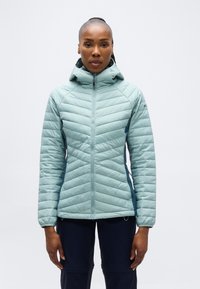 Kvinne vendt fremover iført en lyseblå quiltet hettejakke og mørke bukser mot en enkel hvit bakgrunn.