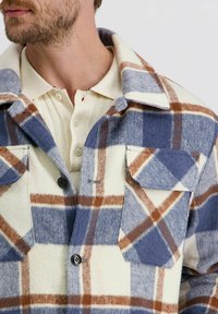 Man in een crème gebreide polo onder een blauw, bruin en crème geruite wollen jas met twee borstzakken en zwarte knopen.