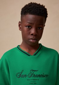 Green sweatshirt with a V-neck and black decorative trim. Features the text "San Francisco California" in dark lettering. Smooth fabric texture.