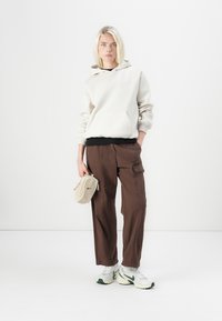 Light beige hoodie with a kangaroo pocket, brown cargo pants, and white sneakers with green accents. Holding a small beige bag.