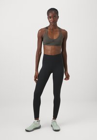 Reggiseno sportivo di colore verde oliva con spalline sottili, abbinato a leggings neri a vita alta. Le calzature sono di un colore verde chiaro con accenti scuri.
