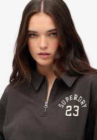 Felpa a mezza zip marrone scuro con colletto, caratterizzata da scritte ricamate bianche "SUPERDRY" e il numero "23" sul petto.