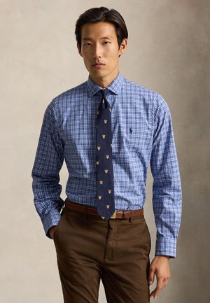 Homme portant une chemise à carreaux bleue, une cravate bleu marine avec des motifs de blason, une ceinture marron et un pantalon marron, debout devant un fond beige.