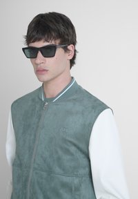 Bomber jacket verde in suede con manica in pelle bianca, chiusura con zip e collare a costine. Occhiali da sole angolari neri completano il look.