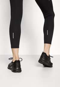 Leggings sportivi neri con dettagli riflettenti, realizzati in un tessuto liscio ed elastico e abbinati a scarpe da corsa nere con suole testurizzate.