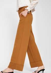 Pantalon large de couleur marron chaud, fabriqué dans un tissu lisse avec des poches latérales et un ourlet roulé. Associé à un blouse en dentelle.