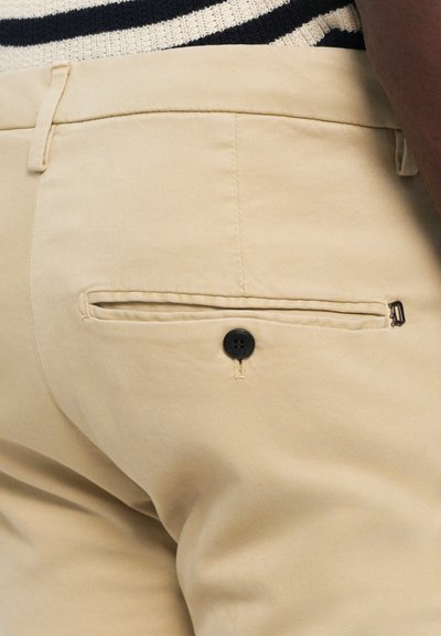 Primo piano di pantaloni beige che mostrano la tasca posteriore con bottone nero e passanti per cintura, indossati con un top in maglia a righe.