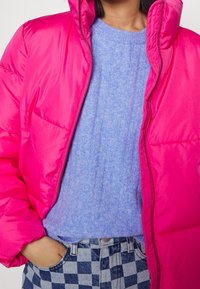 Ljusrosa puffjacka med hög krage och dragkedja, lagd över en ljusblå ribbad tröja, parat med rutiga denimbyxor.