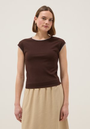 Mujer joven con cabello castaño lacio, vistiendo una blusa sin mangas ajustada de color marrón oscuro y una falda beige, posando frente a un fondo liso.