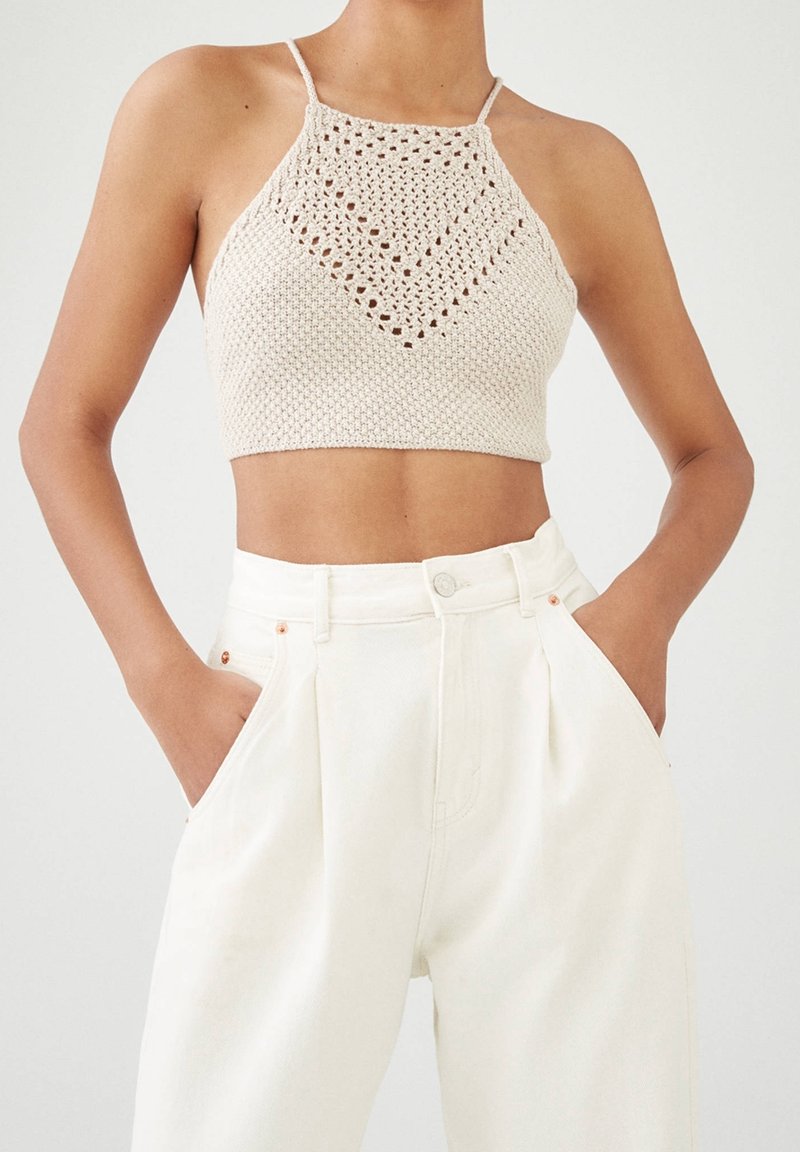 Beżowy crop top na szydełku z ażurowym wzorem w diamenty, w zestawie z białymi spodniami o wysokim stanie, z fałdami i kieszeniami.