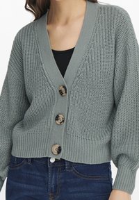 Gilet tricoté vert avec un décolleté en V, trois grands boutons et des poignets et un ourlet côtelés. La texture est épaisse et chaude. Porté par-dessus un top noir.