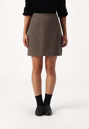 Kvinde iført en brun mininederdel med hundetandmønster, sorte sokker, sorte loafers og en sort top med puffede ærmer, stående på hvidt gulv.
